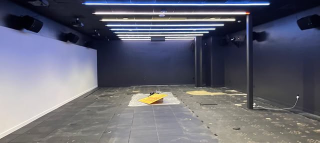 An empty room with black walls and ceiling, featuring linear ceiling lights and a partially removed black floor with exposed patches.