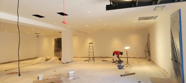 A room under construction with a person working, a ladder, scattered tools, and materials on the floor. The ceiling is partially unfinished, and there are exposed wires hanging down.
