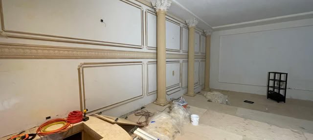 A room under renovation with ornate wall panels, two columns, construction materials on the floor, and a small black shelving unit against the wall.