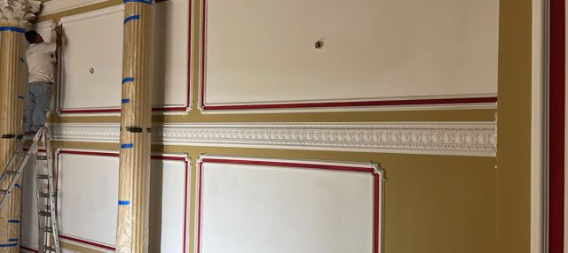A person on a ladder working on a decorative wall with columns and red-bordered panels.